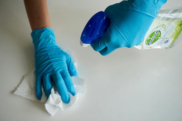 An individual in blue gloves is wiping a table with kitchen towel and multi-surface cleaner
