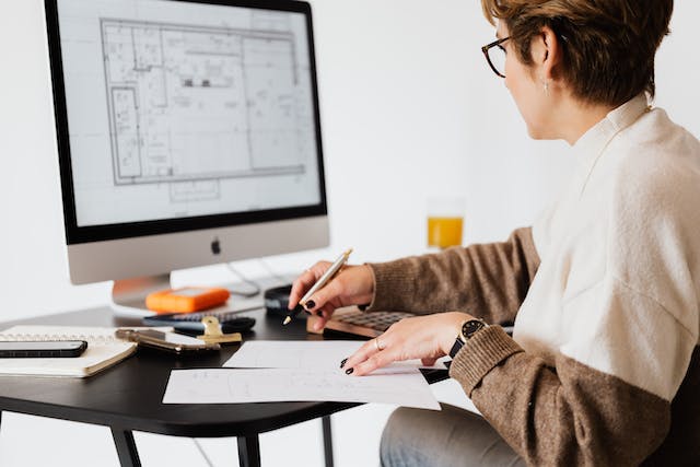 A woman, seated at a desk, reviews a refinishing project plan on her computer while writing on a piece of paper