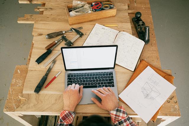 A person sits at a wooden table, using a laptop to design for a refinishing project