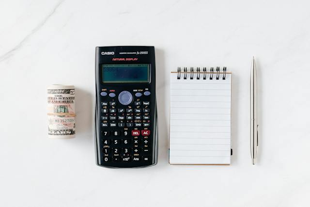 A calculator, notepad, pen, and a bundle of dollars neatly arranged on a pristine white marble surface